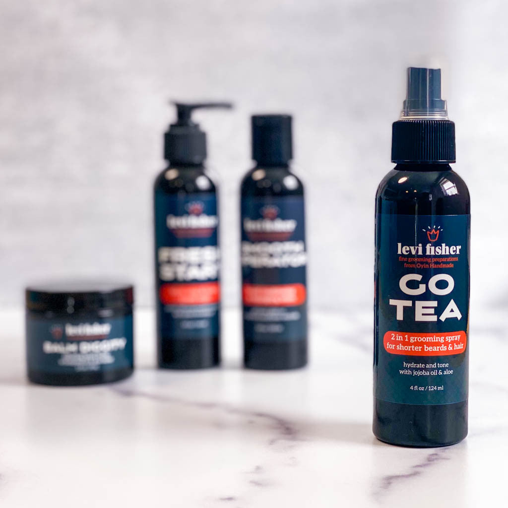 Go tea hydrating beard spray is pictured in a black bottle on marble countertop. Other products in this collection appear blurry in the background.