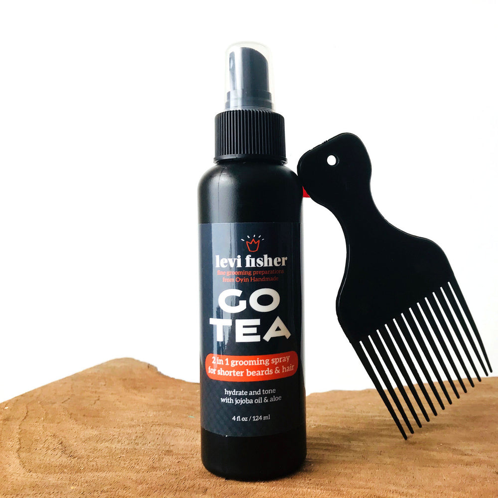 Go tea hydrating beard spray is pictured in a black bottle on a wooden cutting board. An afro-pick leans against the bottle.
