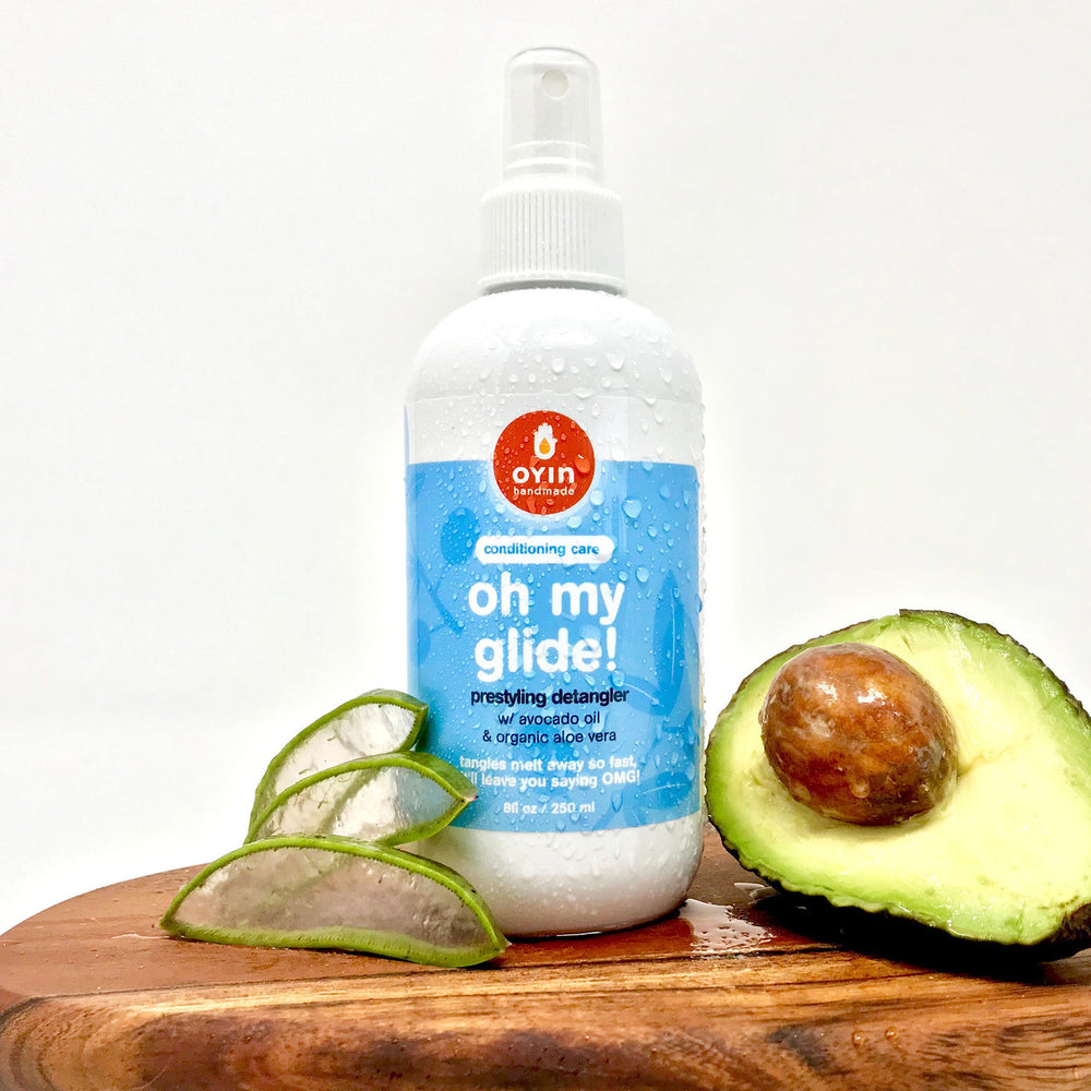 8oz bottle of Oh My Glide detangler in a spray bottle, standing up on a wooden cutting board between some aloe vera plant slices and a halved avocado. 