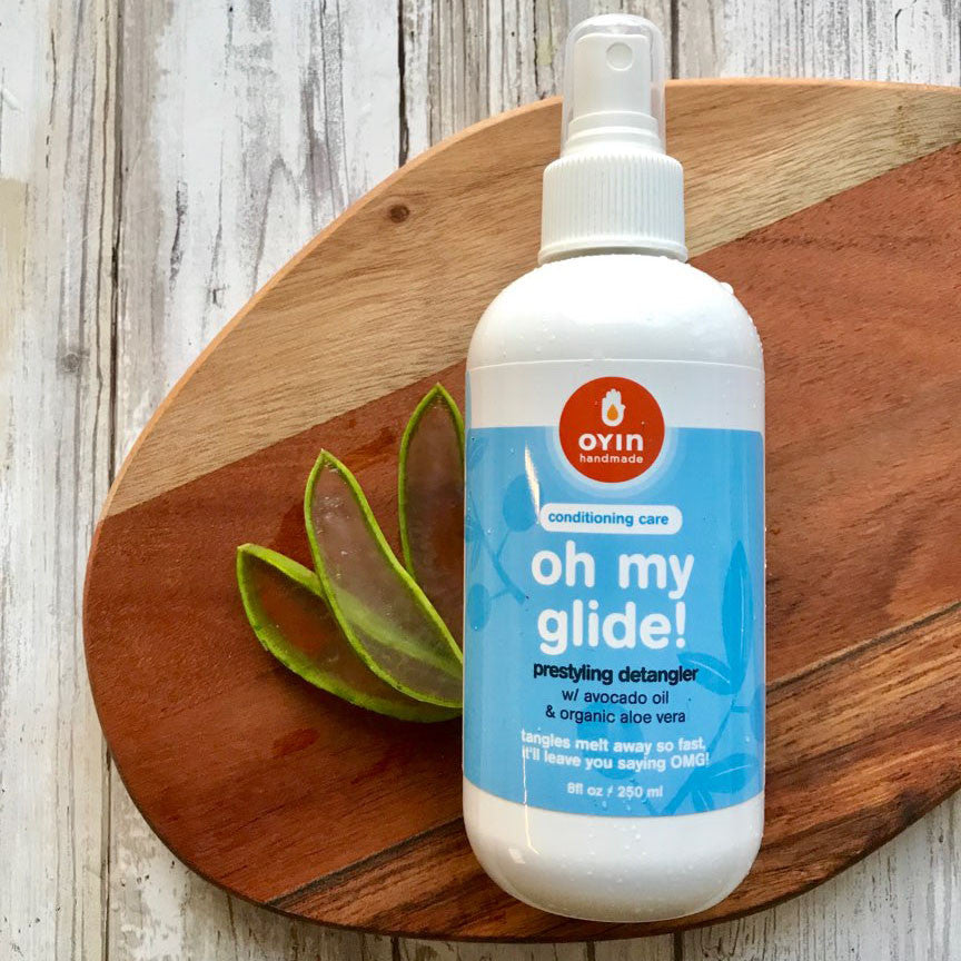 8oz bottle of Oh My Glide detangler in a spray bottle, laying flat on a wooden cutting board with aloe vera plant slices next to it.